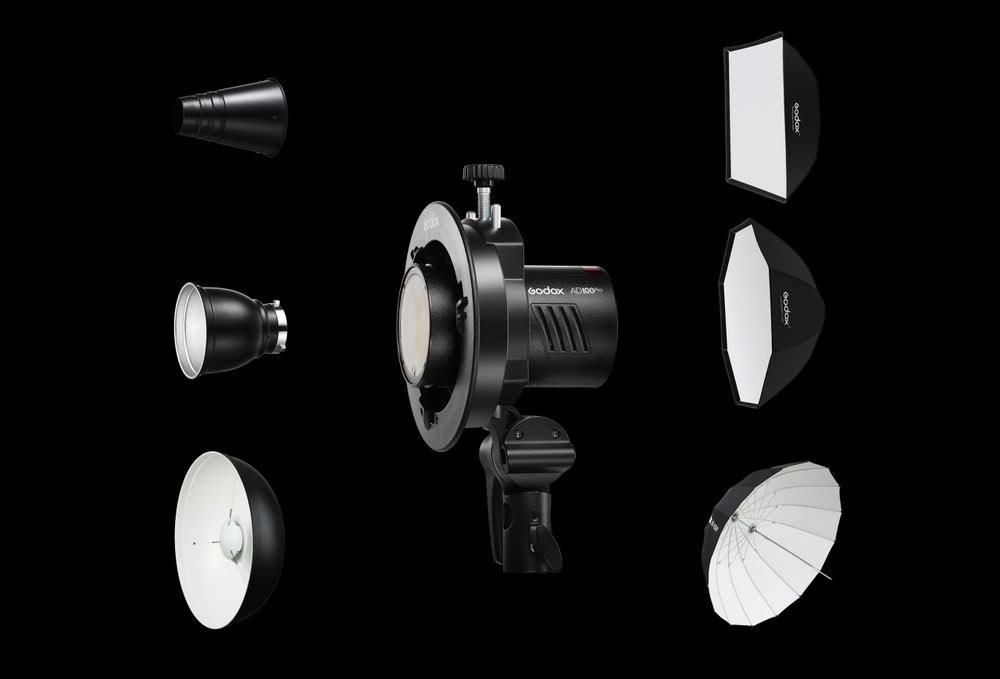 Акумуляторний спалах Godox AD100Pro в центрі, оточений сумісними модифікаторами світла на чорному фоні: тубус, рефлектор, біла портретна тарілка, софтбокс, октобокс та парасолька на просвіт